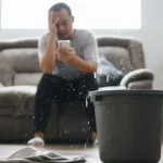 A man sitting on the sofa with a phone in hand and a stressed look on his face. In the foreground, a ceiling leaking into a bucket.